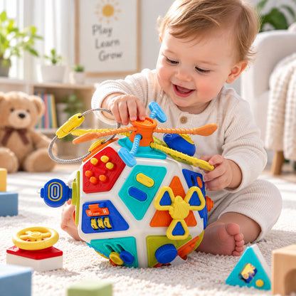 A baby sitting on a carpeted floor in a bright nursery, smiling and reaching for a colorful, spherical activity toy with multiple interactive panels and a helicopter attachment. The baby is wearing a cream-colored ribbed onesie, and a teddy bear and framed sign are visible in the softly lit background.
